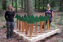 Zwei Frauen stehen im Wald neben einer Versuchsanlage mit selbtgebauten Bäumchen aus Holz